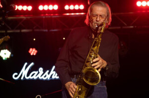 Terry Marshall, founder of Marshall Amplification playing saxophone on stage. A neon Marshall logo is alight behind him.